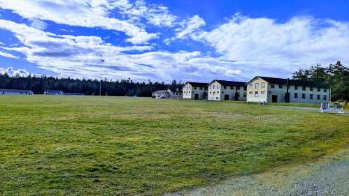 A grassy field with several buildings under a blue sky with clouds, surrounded by trees in the background.