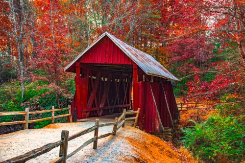 A red covered bridge surrounded by vibrant autumn foliage and a gravel path, framed by wooden fences.