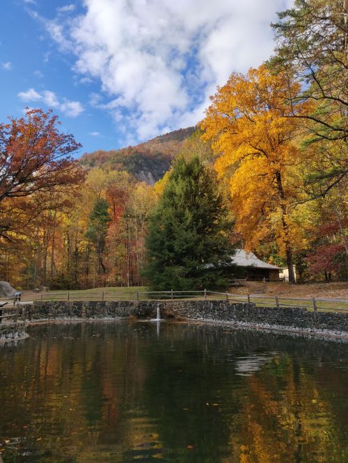 A serene pond reflects vibrant autumn trees and a blue sky, with mountains in the background.