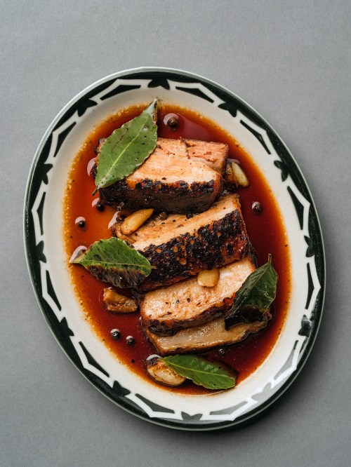 Sliced, glazed pork belly on a decorative plate, garnished with bay leaves and garlic in a rich sauce.