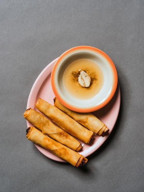 A plate with crispy spring rolls and a small bowl of dipping sauce, featuring a slice of garlic on top.