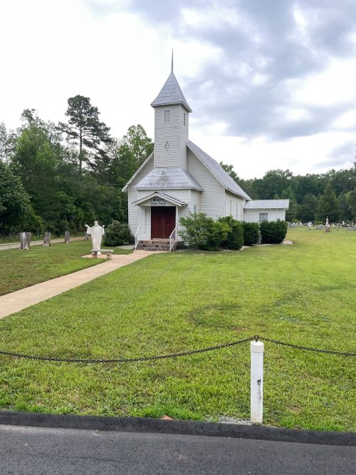 A small white church with a steeple, surrounded by green grass and trees, near a cemetery. Overcast sky above.
