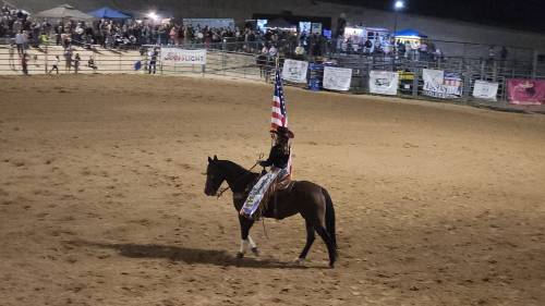 A rider on horseback carries the American flag in a rodeo arena, with a crowd and banners in the background.