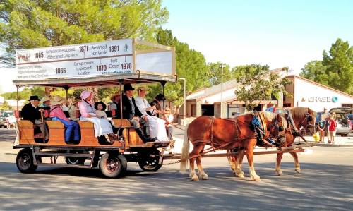 A horse-drawn carriage with passengers in historical attire rides through a town, with shops in the background.