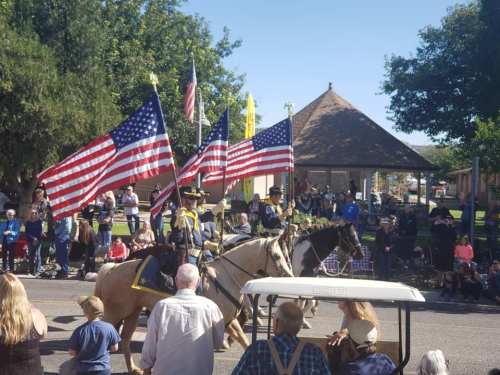 A parade scene with riders on horseback carrying American flags, surrounded by a crowd and trees in the background.
