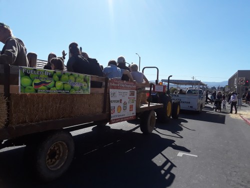 A hayride with people sitting on bales, passing by a truck on a sunny day. Signs on the wagon promote local businesses.