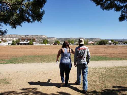 Two people walking away on a dirt path, with a scenic landscape and blue sky in the background.
