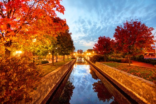 A serene canal lined with colorful autumn trees, reflecting lights and clouds in the water at dusk.