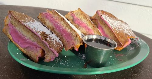 A plate with four slices of stuffed French toast filled with pink cream, dusted with powdered sugar, and a small cup of syrup.