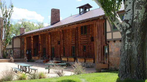 A rustic log cabin building with a stone foundation, surrounded by green grass and trees under a clear blue sky.