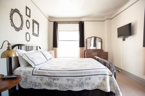 A cozy bedroom featuring a bed, mirrors on the wall, a dresser, and a TV, with natural light coming through the window.