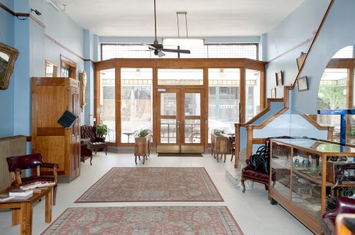 A spacious, well-lit lobby with blue walls, wooden furniture, and decorative rugs, featuring large windows and a staircase.