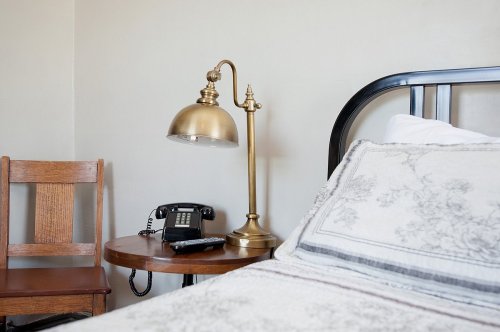 A cozy bedroom corner featuring a bed, a brass lamp, a vintage phone, and a wooden chair.