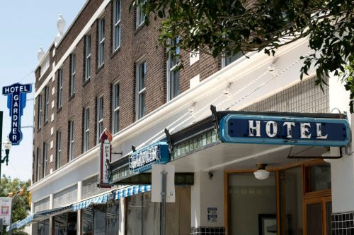 Exterior of the Hotel Gardner, featuring a vintage blue sign and brick facade, with nearby shops visible.
