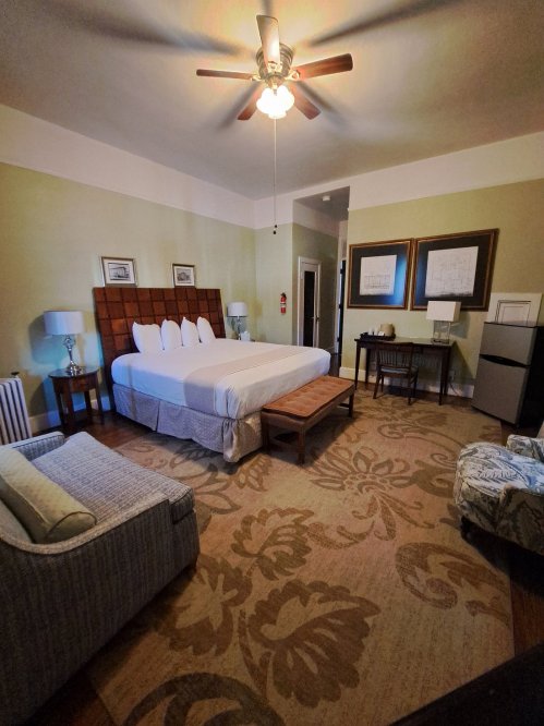 Cozy hotel room featuring a large bed, seating area, desk, and decorative rug. Warm lighting and soft colors create a welcoming atmosphere.