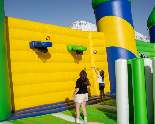 Two children play on a colorful inflatable structure with basketball hoops against a backdrop of mountains.
