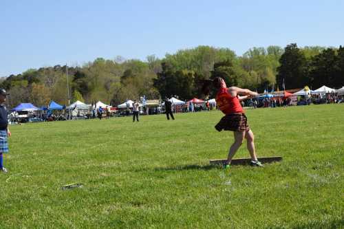 A woman in a red top and kilt throws a heavy object in a grassy field during a festival, with tents in the background.