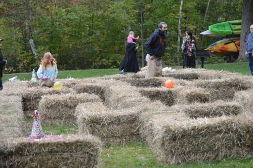 A group of people play in a maze made of hay bales, with colorful balloons scattered around in a wooded area.