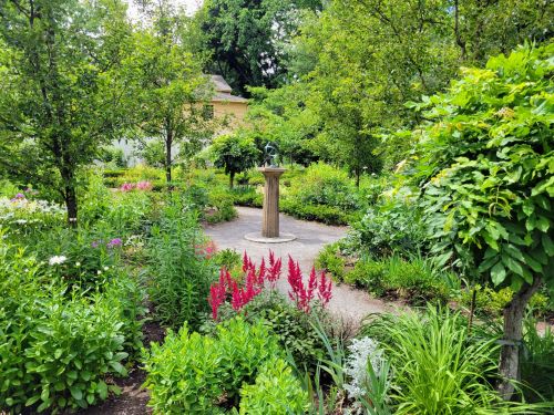 A lush garden with vibrant flowers, greenery, and a central stone pedestal surrounded by trees.