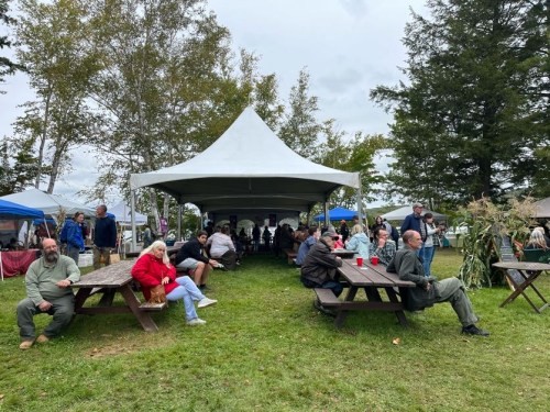 A gathering under a large tent with people sitting at picnic tables, enjoying the outdoors and socializing.