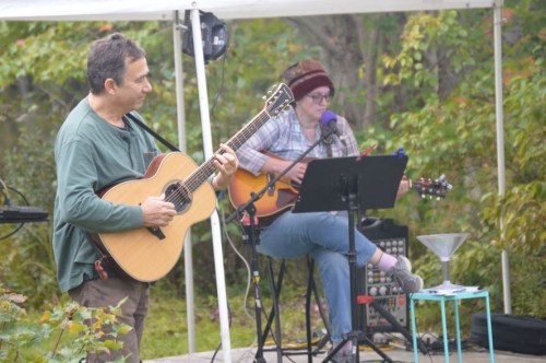 Two musicians perform outdoors under a tent, one playing guitar and the other singing, surrounded by greenery.