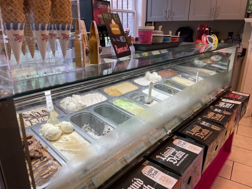 A display case filled with various ice cream flavors and toppings, with cones and cups in the background.