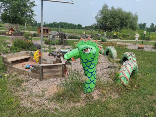 A colorful dragon sculpture in a grassy play area, surrounded by wooden play structures and greenery.