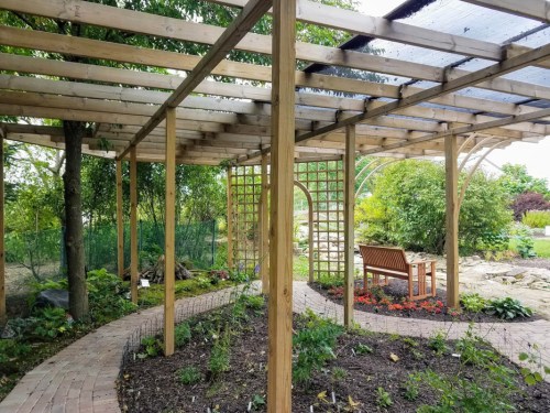A wooden pergola with a bench surrounded by greenery and flower beds in a serene garden setting.