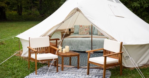 A cozy glamping tent with a bed inside, two wooden chairs, a small table, and candles on a patterned rug outside.