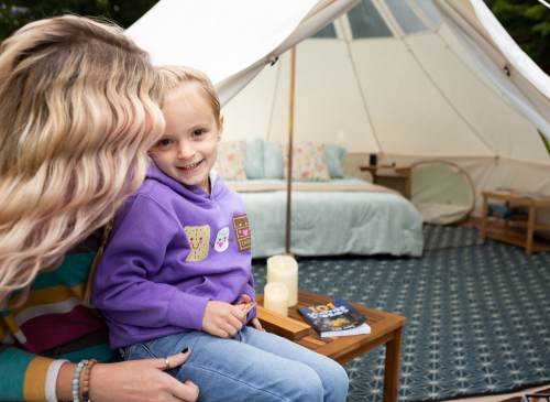 A woman and a young boy smile together outside a cozy tent, with a bed and decor visible inside.