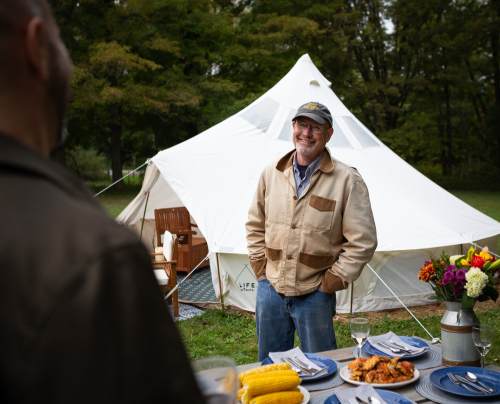 A smiling man in a jacket stands near a dining setup outdoors, with a tent and nature in the background.