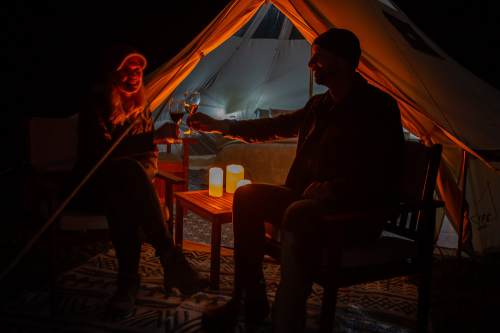 A couple toasts with glasses of wine by a glowing tent at night, surrounded by candles and a cozy atmosphere.