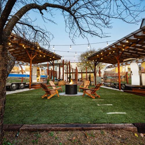 A cozy outdoor space with vintage Airstreams, wooden seating, and string lights, centered around a fire pit.