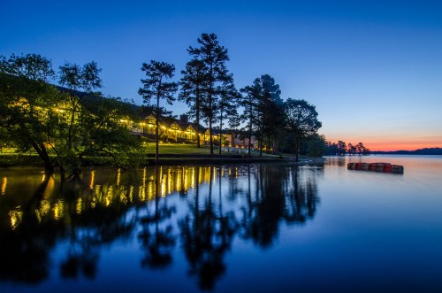 A serene lakeside scene at dusk, with reflections of trees and lights on the water, and a colorful sunset in the background.