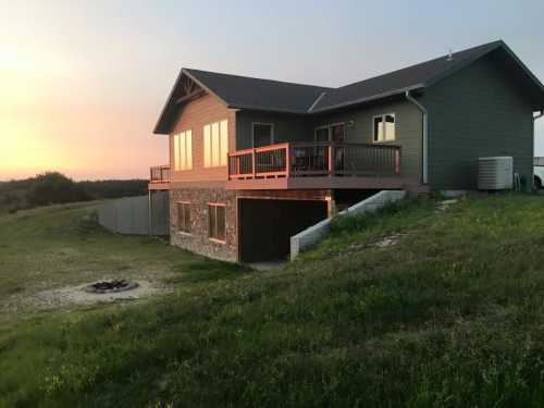 A modern house with a deck, set on a grassy hill, overlooking a fire pit at sunset.