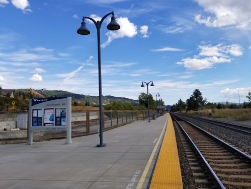 A train station platform with yellow markings, lamps, and a schedule board, surrounded by trees and mountains under a blue sky.