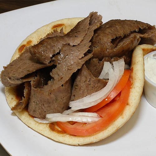 A plate with pita bread filled with sliced meat, tomatoes, onions, and a side of sauce.