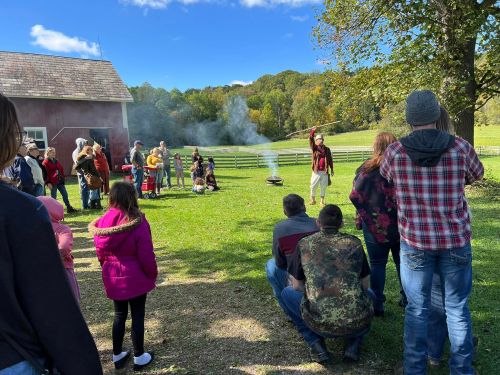 A group of people watches a demonstration outdoors, with smoke rising from a fire pit and a scenic background of trees.