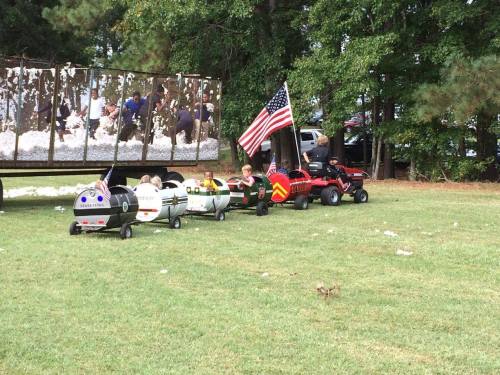 Children riding small toy cars in a line, with a backdrop of a trailer and an American flag nearby.