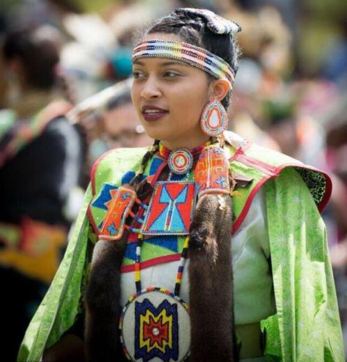 A young woman in traditional attire with colorful beading and accessories, smiling at a cultural event.