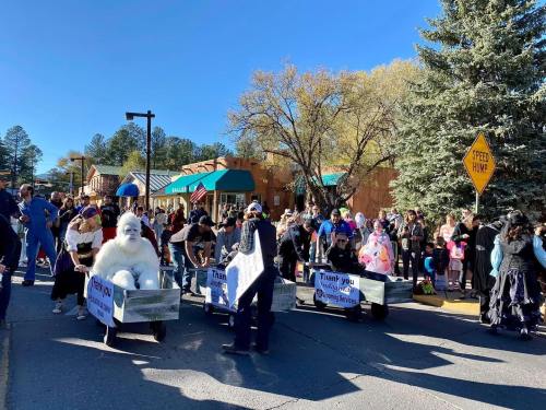 A festive parade scene with people in costumes, including a yeti, walking alongside decorated wagons on a sunny day.