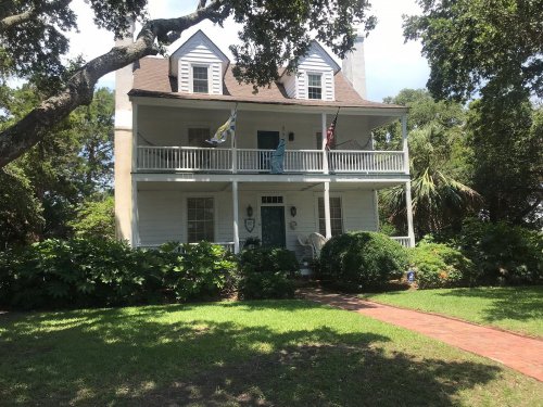 A two-story white house with a brown roof, surrounded by greenery and featuring a front porch with flags.