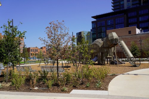 A playground with a slide and climbing structure, surrounded by trees and benches, under a clear blue sky.