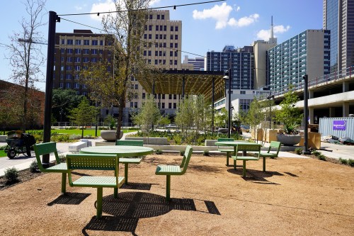 A sunny urban park with green tables and chairs, surrounded by buildings and trees under a clear blue sky.