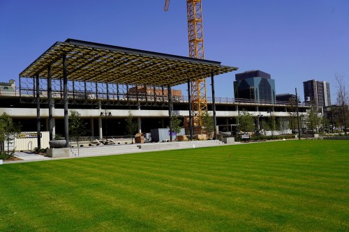 A modern outdoor space with a large canopy, green lawn, and construction in the background under a clear blue sky.