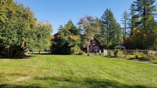 A sunny landscape featuring a red house, green lawn, trees, and people walking in a peaceful outdoor setting.
