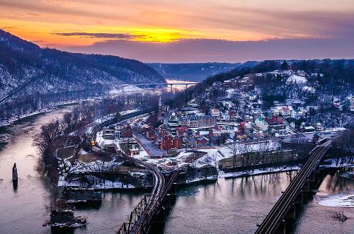 A scenic view of a river town at sunset, surrounded by hills and snow, with bridges crossing the water.