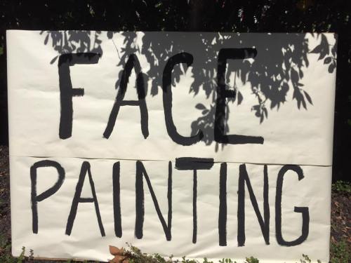 A large sign reading "FACE PAINTING" in bold black letters on a white background, surrounded by greenery.