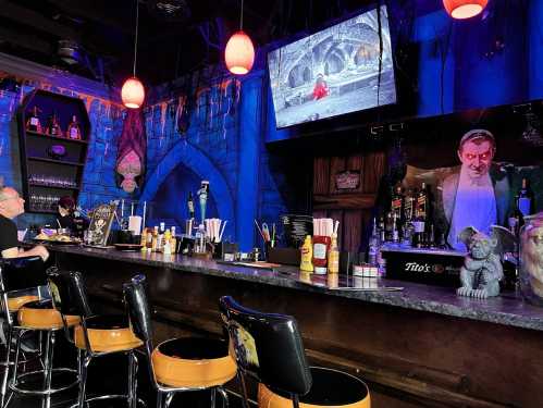 A spooky bar with blue walls, a TV showing a horror scene, and Halloween decorations. Bar stools are lined up at the counter.