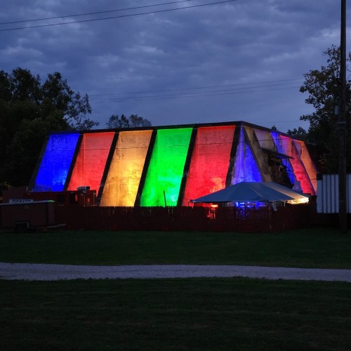 A colorful, illuminated building with red, green, and blue lights against a cloudy sky, surrounded by grass and a fence.